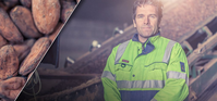 A white, brown-haired man in a warehouse jacket. He's standing in a warehouse. The background is blurry, but you can see cocoa on a conveyor belt. The man is facing the camera, he looks determined and happy.