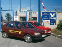 Two red cars in front of a gate. The cars have Berthold Vollers written on them and are therefore company cars. On a wall next to the gate, signs can be seen. They aren't completely clear readable, but it is obvious from them that we are at the Vollers location in Riga.