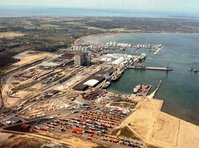 The photo is taken from above. Pictured is a port. There are lots of containers in the front as well as buildings. Water and green areas can be seen in the background.
