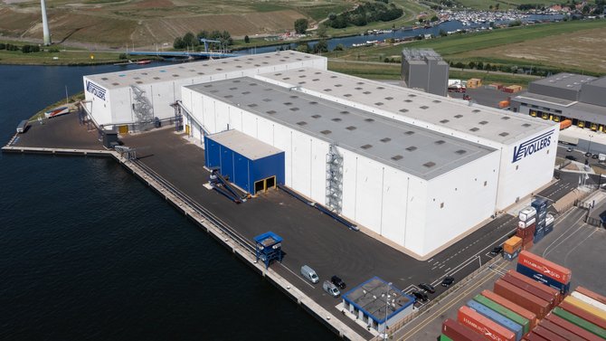 A photo taken from above. Centered are three white warehouses with a blue logo on the walls. It reads "Vollers". Left to the warehouses and behind it, is water. To the right, containers in all colours can be seen.