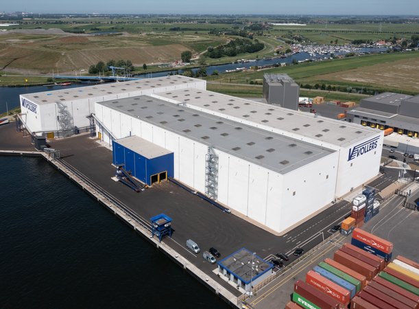 [Translate to Nederlands:] A photo taken from above. Centered are three white warehouses with a blue logo on the walls. It reads "Vollers". Left to the warehouses and behind it, is water. To the right, containers in all colours can be seen.