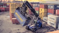 Blue reachstacker lifting a container into the air. It is standing in a harbour, around it are other containers.