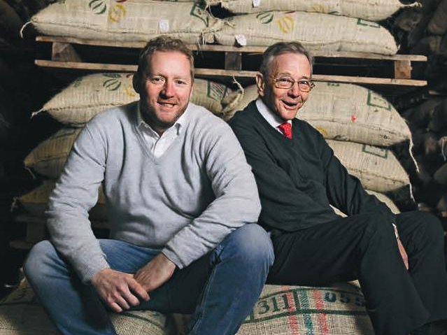 Two white men are sitting on a staple of coffee bags. The man on the left has blonde to red hair and wears a white dressing shirt and a grey sweater with a pair of blue jeans and black shoes. It's Christian Vollers. Next to him sits his father, Lüder Vollers. He's got grey hair and wears glasses, a white dressing shirt with a red tie, combined with a dark green sweater, black trousers and black shoes. They are both smiling at the camera.