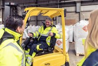 Three white people in a warehouse talking to each other. A blonde woman is seen from behind, a man is smiling at her. The third person is a man in a forklift, he's smiling too. 