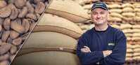 A white man with a dark blue cap and sweater in a warehouse. He's leaning against some coffee bags, his arms are crossed. He's facing the camera, smiling.